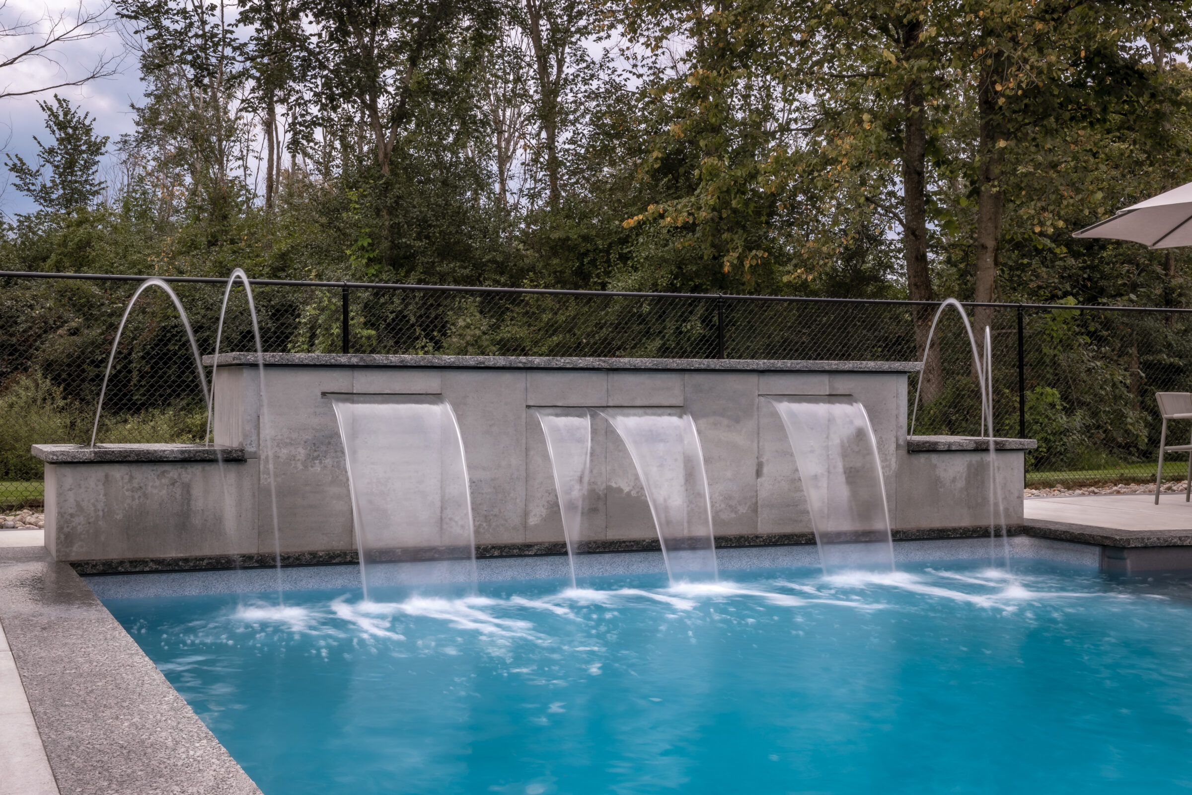 A modern swimming pool features water fountains against a backdrop of lush trees and a gray sky, creating a serene and refreshing atmosphere.
