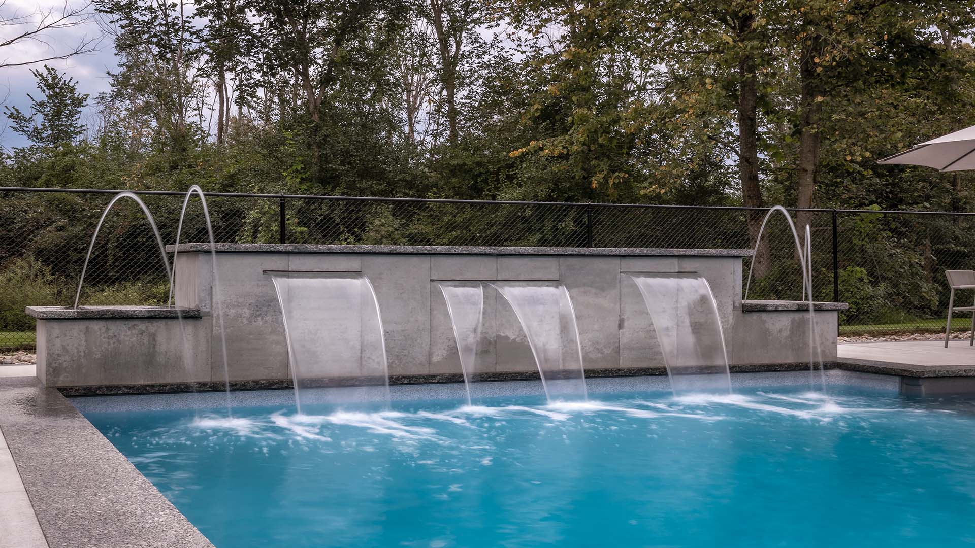 A serene outdoor pool with elegant arching fountains, surrounded by lush greenery and trees, reflecting a peaceful, natural setting. No people present.