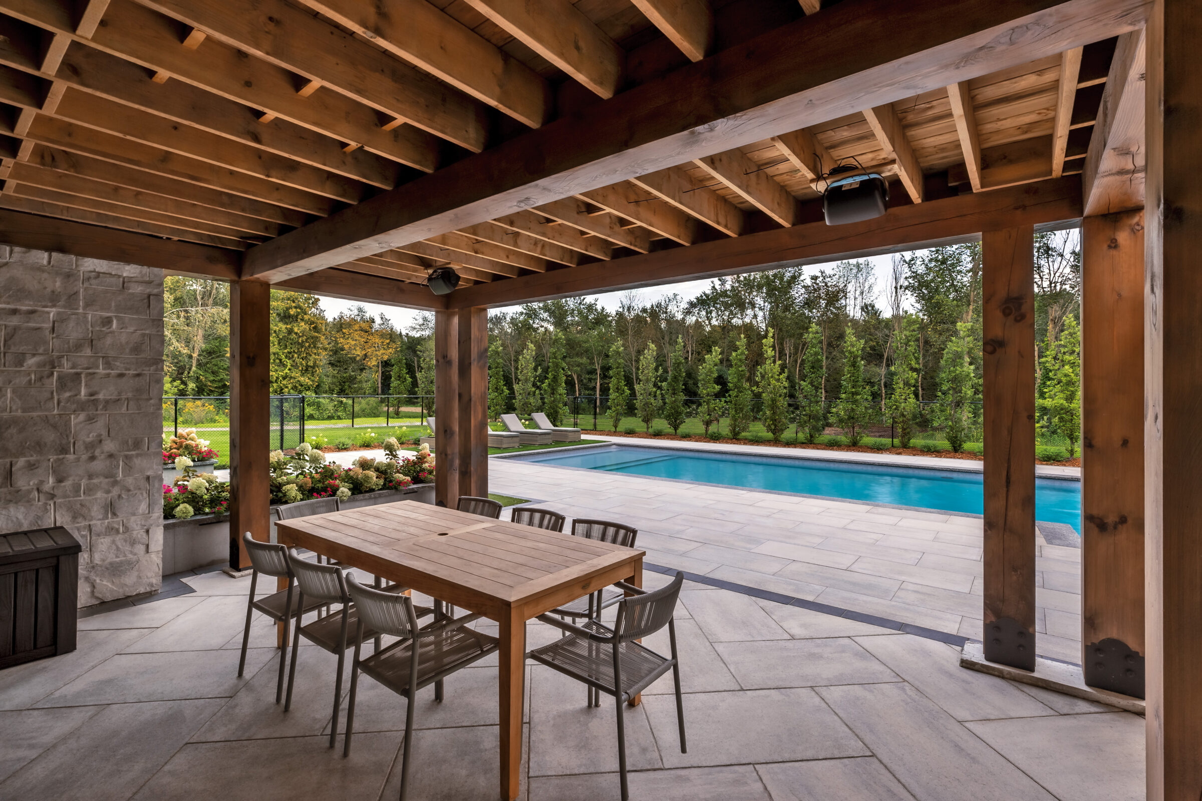 Covered patio with wooden beams, outdoor table and chairs, overlooking a landscaped backyard with a rectangular swimming pool and surrounding trees.