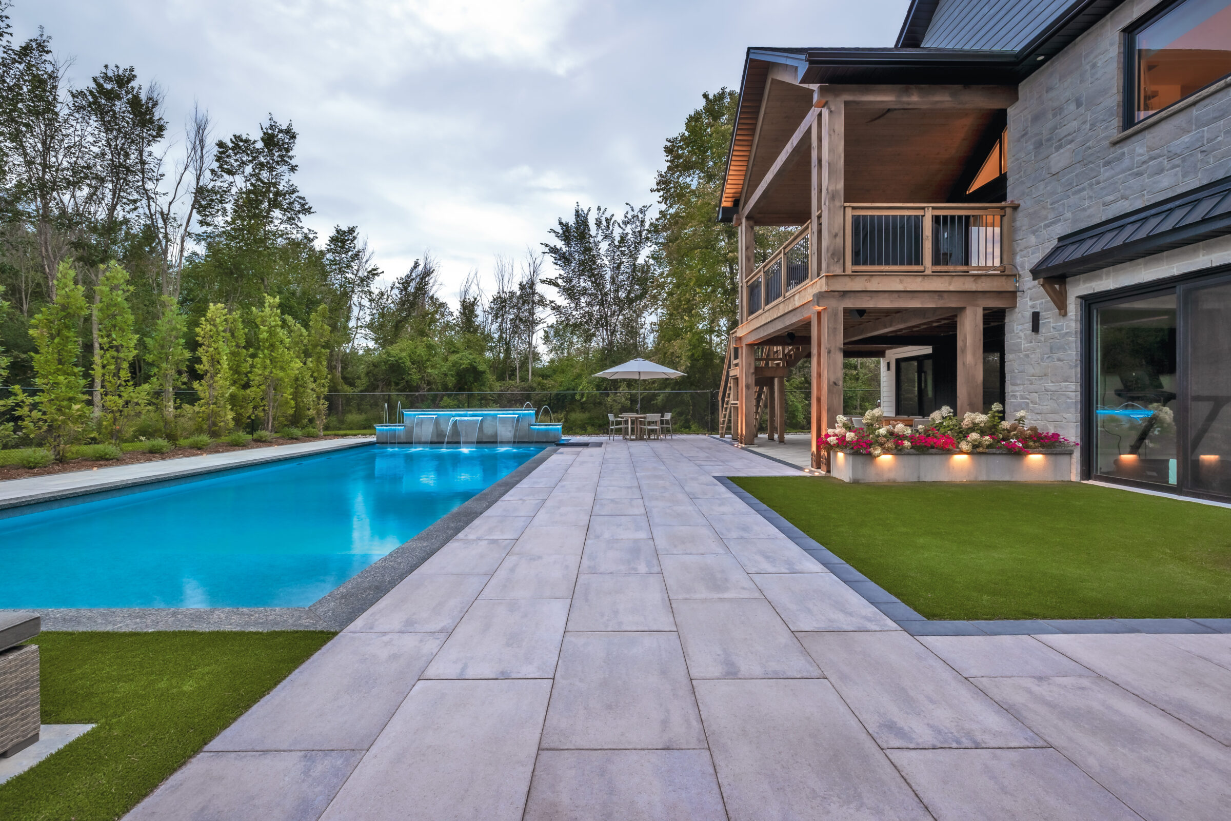 Modern house with spacious patio, pool, and manicured garden. Elevated deck and lush trees create a serene outdoor living space. Cloudy sky overhead.