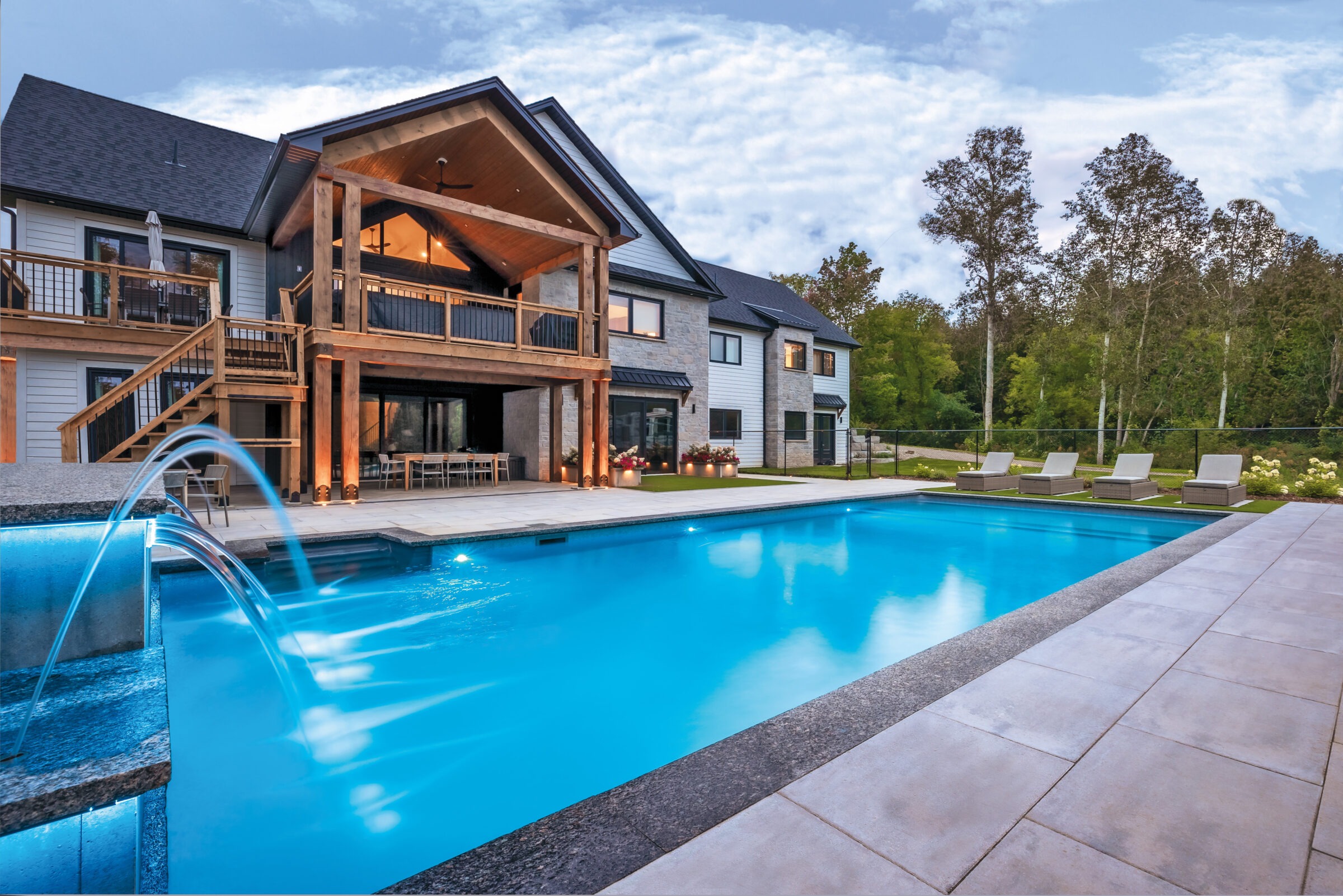 Modern house with large windows, wooden deck, and a spacious backyard featuring a clear swimming pool. Surrounded by trees and patio seating.