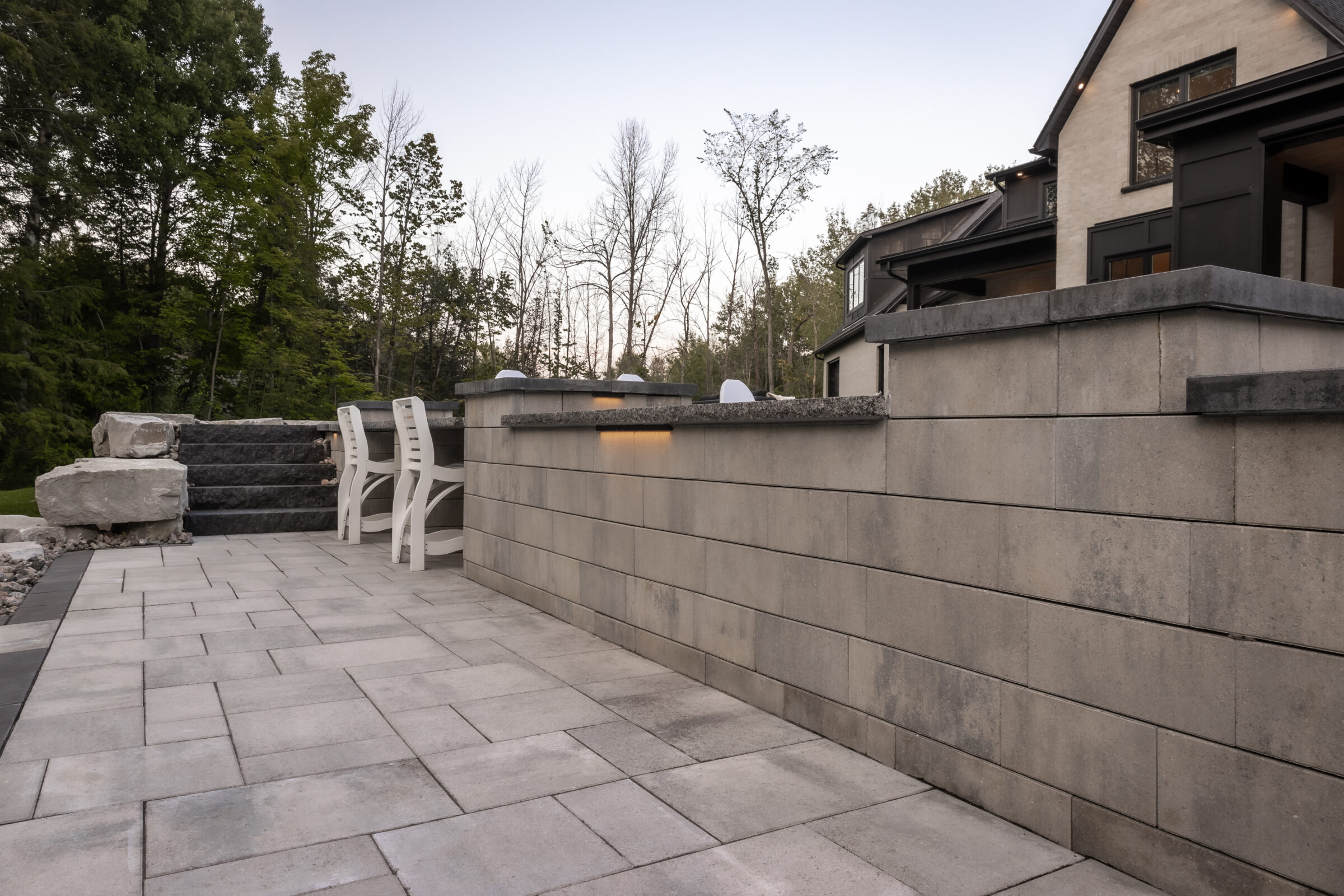 Modern patio with stone tiles, outdoor seating, and stairs. Surrounded by trees, adjacent to a house. Calm, natural setting, no landmarks present.