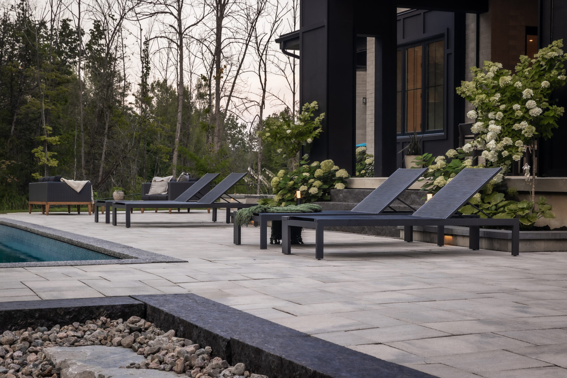 Modern outdoor patio with lounge chairs beside a swimming pool, surrounded by greenery and trees, creating a serene and stylish atmosphere.