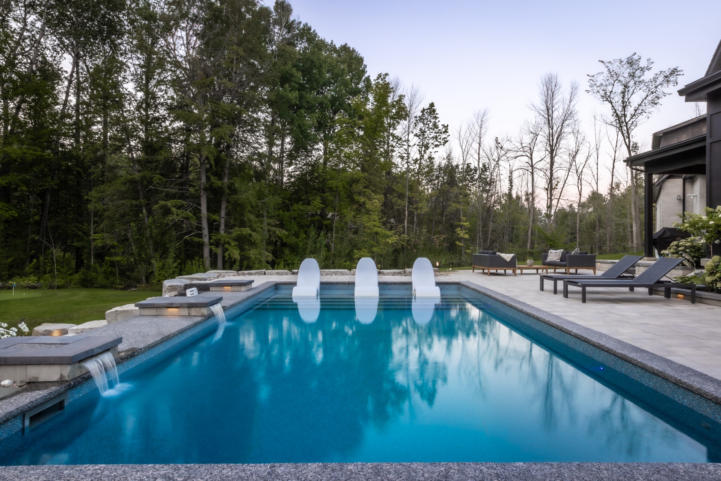 A tranquil backyard features a luxurious swimming pool surrounded by lounge chairs, nestled against a backdrop of lush green trees at dusk.