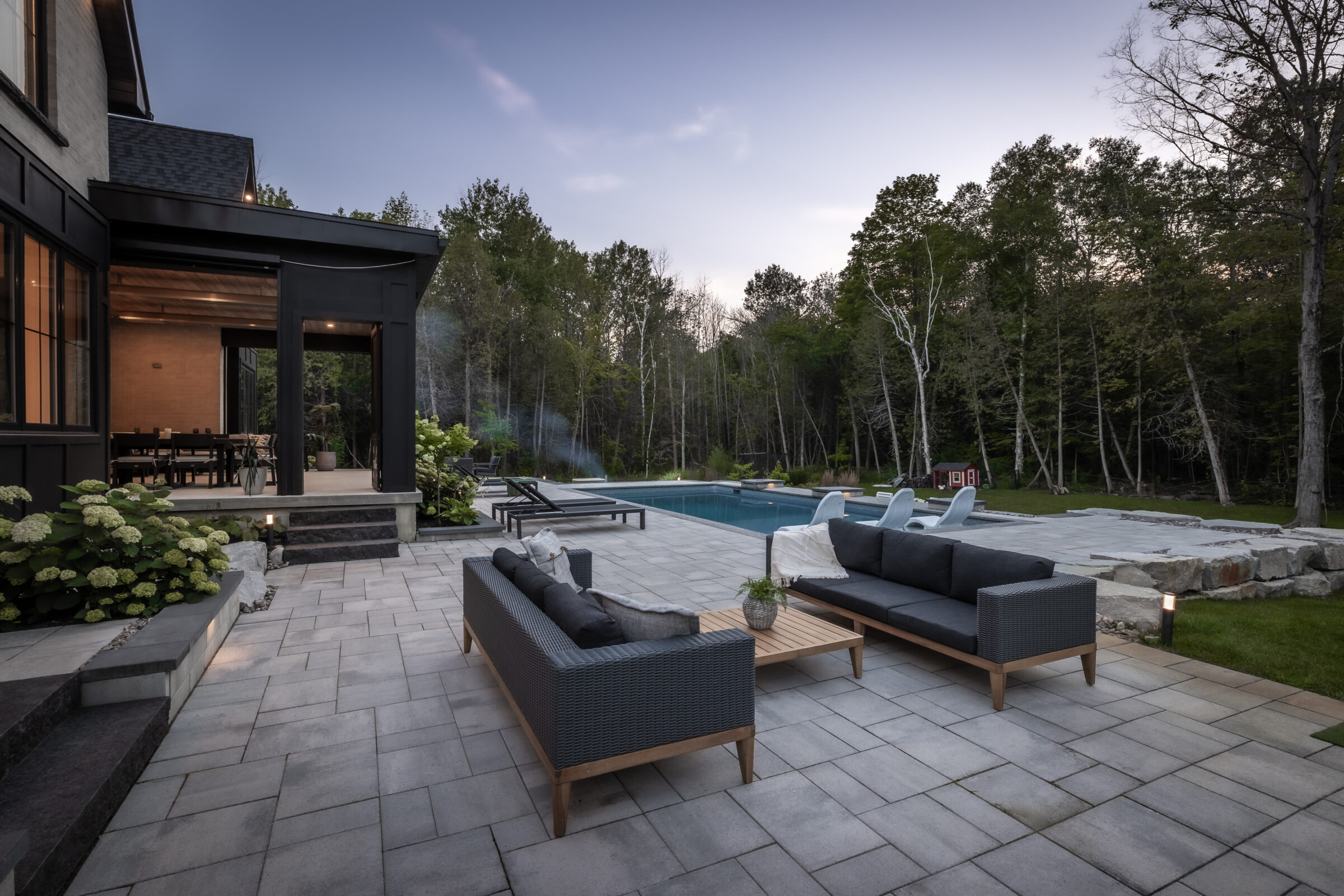 Modern outdoor patio with stylish seating overlooks a tranquil pool surrounded by lush trees. Evening light creates a serene, inviting atmosphere.