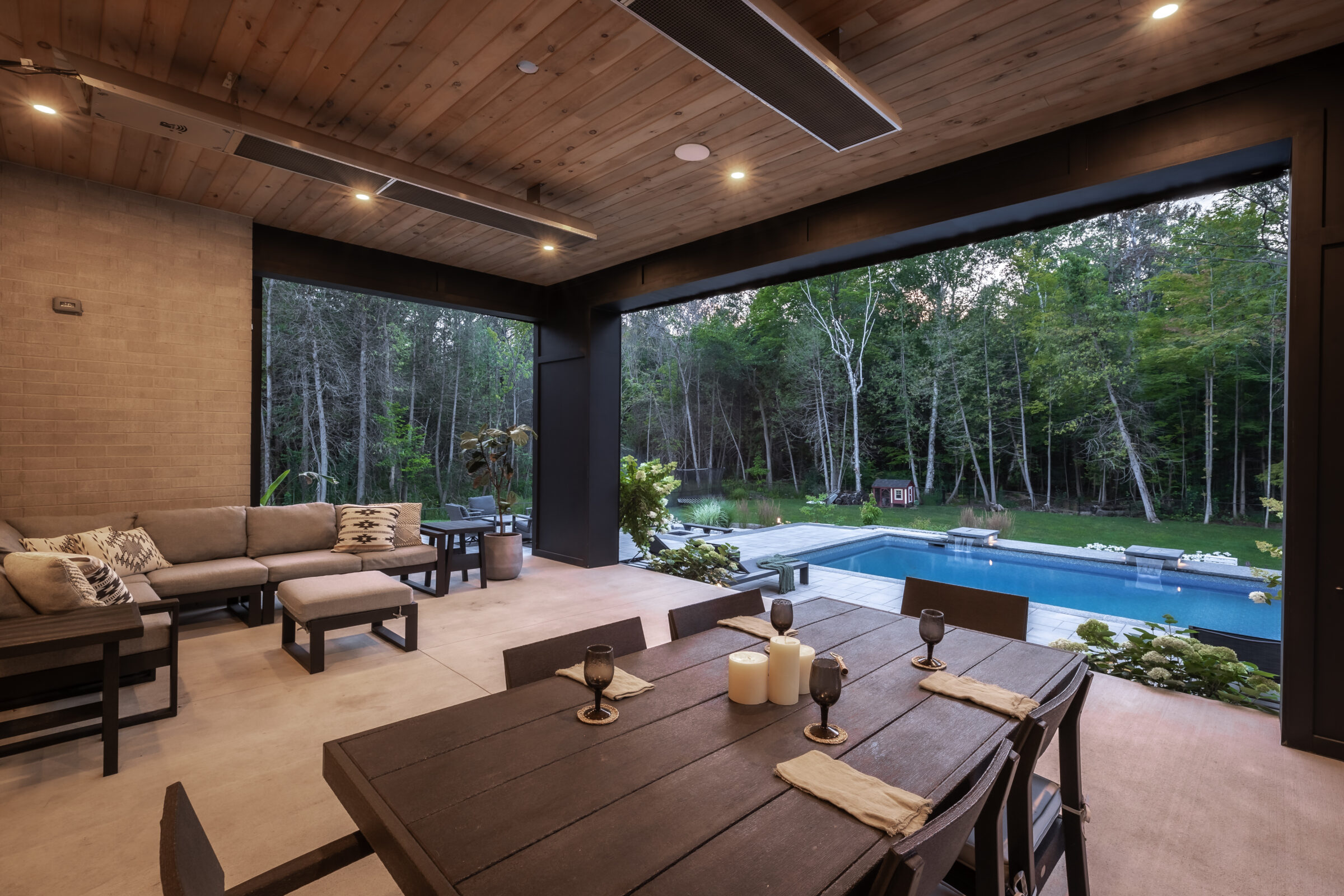 Covered patio with wooden ceiling overlooks a serene pool and lush forest. Modern outdoor seating and dining area create a cozy atmosphere.