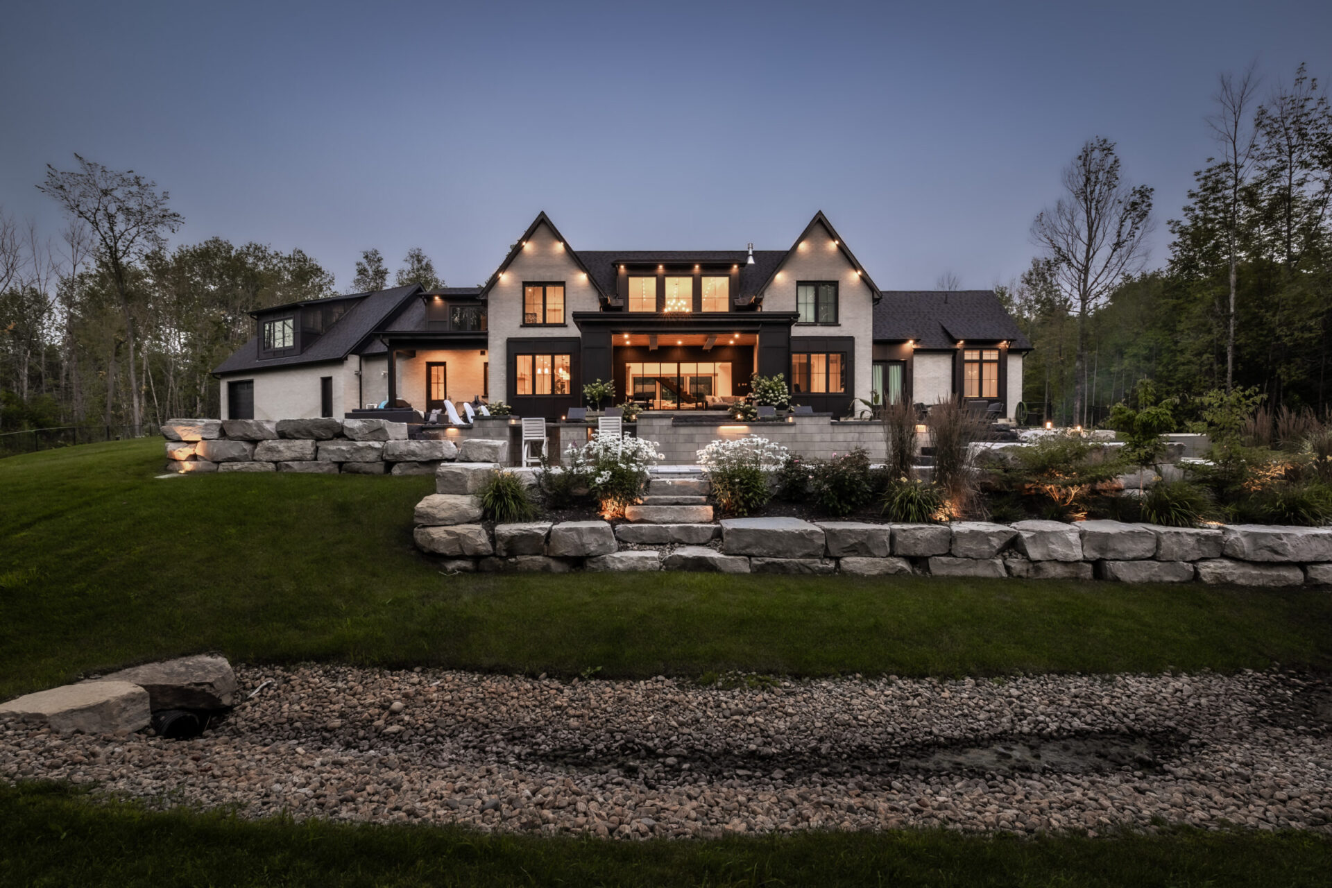 A large, modern house set in a landscaped yard with stone features and outdoor lighting, surrounded by trees and a grassy area.