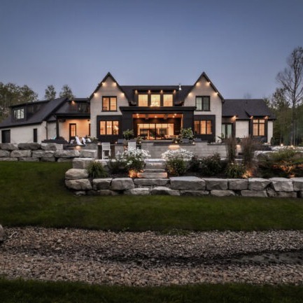 A large modern house with illuminated windows, surrounded by landscaped greenery, rocks, and a stone pathway under a dimly lit evening sky.