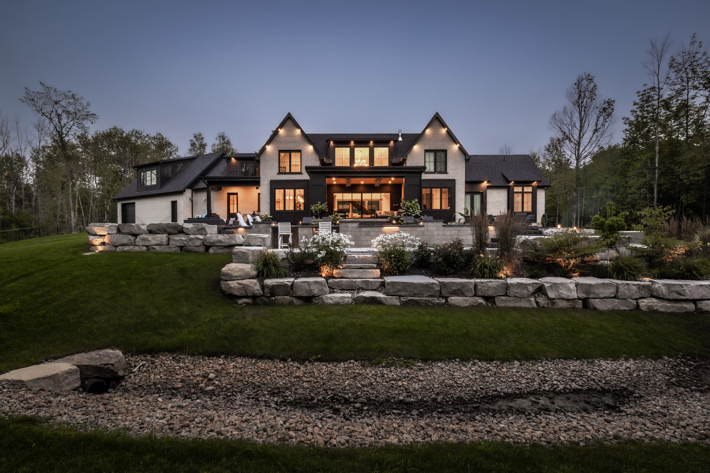A large, modern house set in a landscaped yard with stone features and outdoor lighting, surrounded by trees and a grassy area.