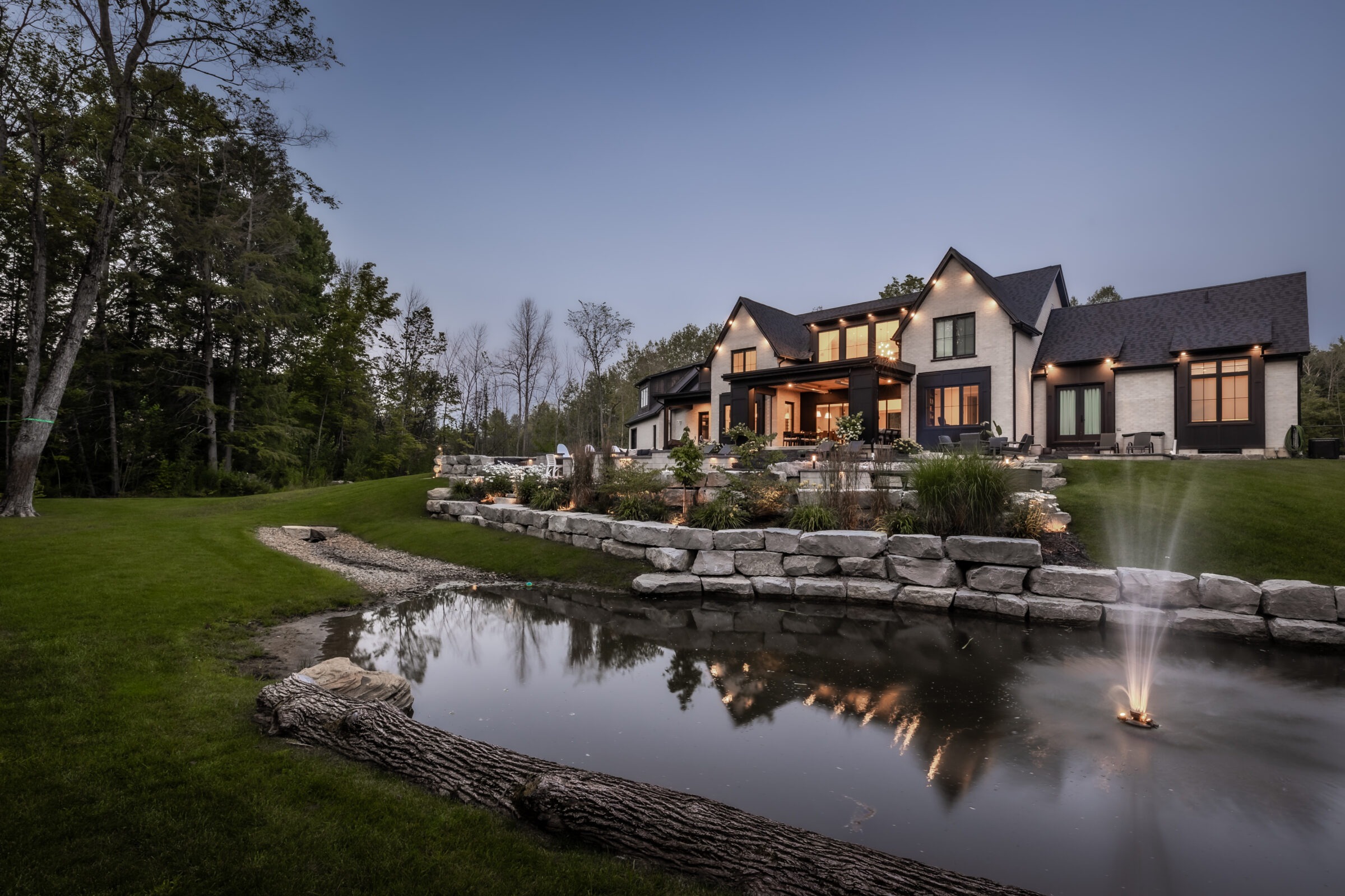 Modern house with large windows, surrounded by trees and manicured lawn, features a small pond with a fountain in the foreground.