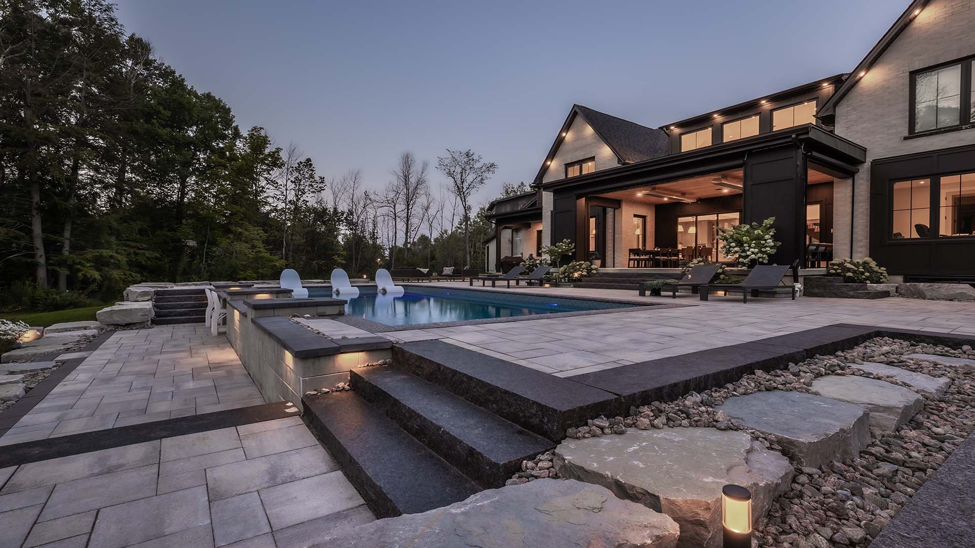 Modern house with large pool and patio, surrounded by trees. Evening lighting creates a serene ambiance. No recognizable landmarks or historical buildings visible.