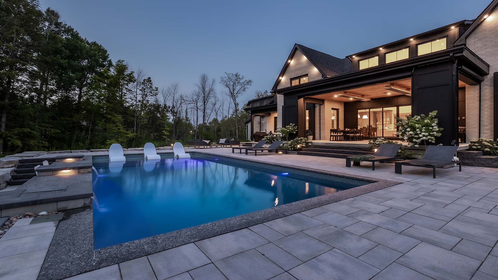 Modern house with large windows, illuminated at dusk. A sleek pool with lounge chairs in the foreground, surrounded by trees and spacious patio.