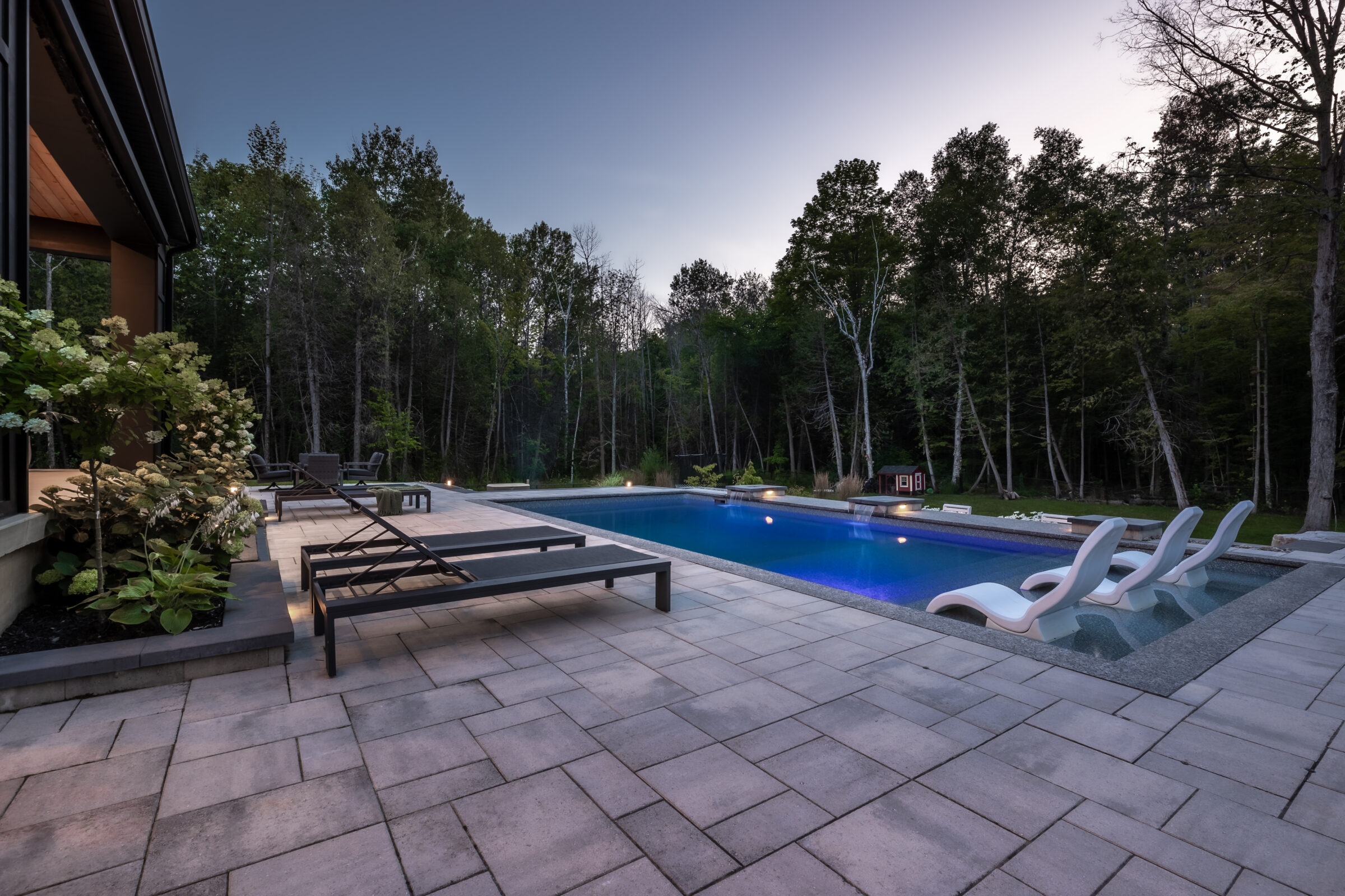 A serene outdoor pool area surrounded by trees, featuring lounge chairs and soft evening lighting, set on a spacious stone patio.