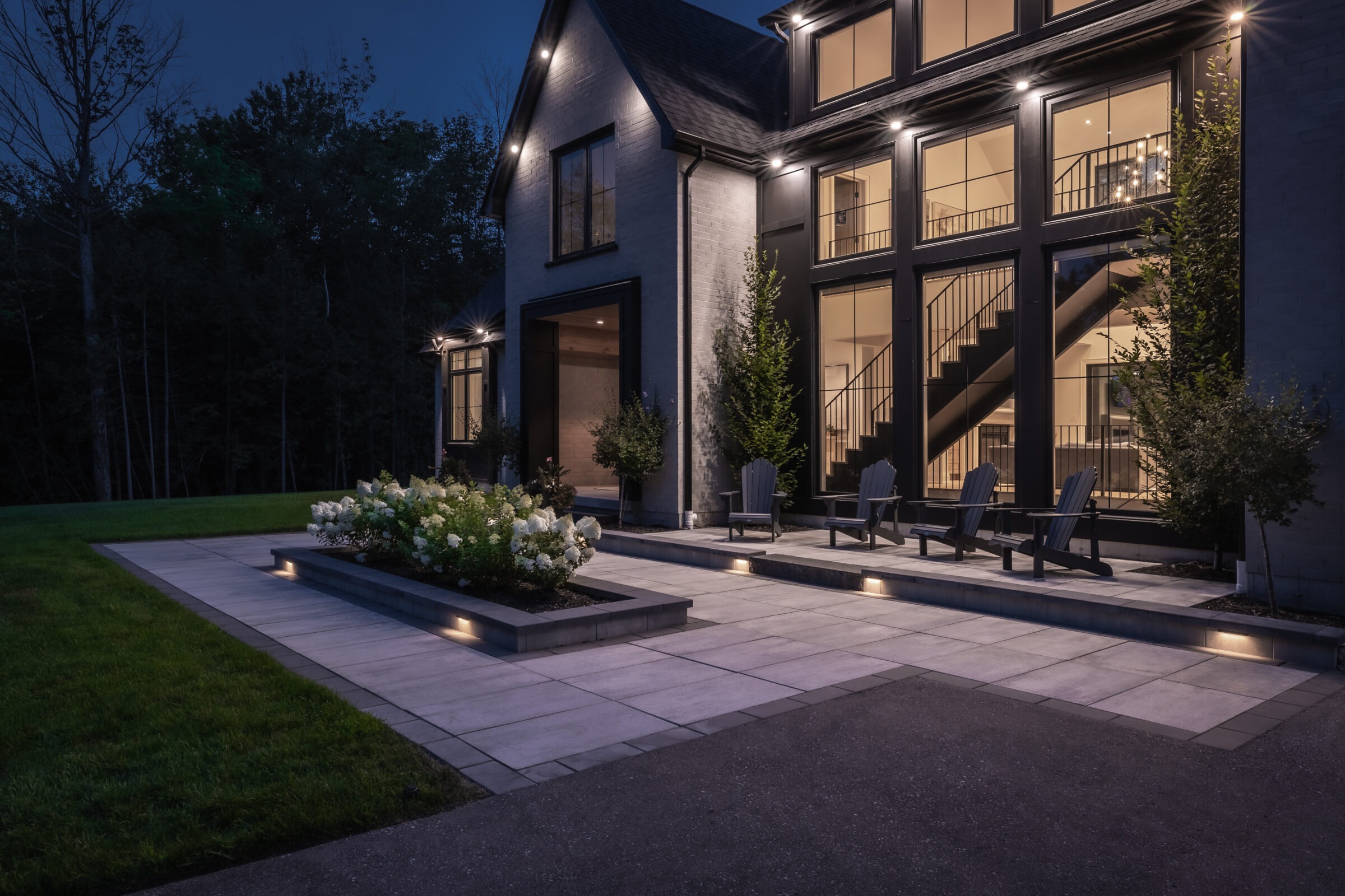 Modern house exterior with large windows and patio, surrounded by plants. Evening setting with subtle lighting. No people or landmarks are visible.