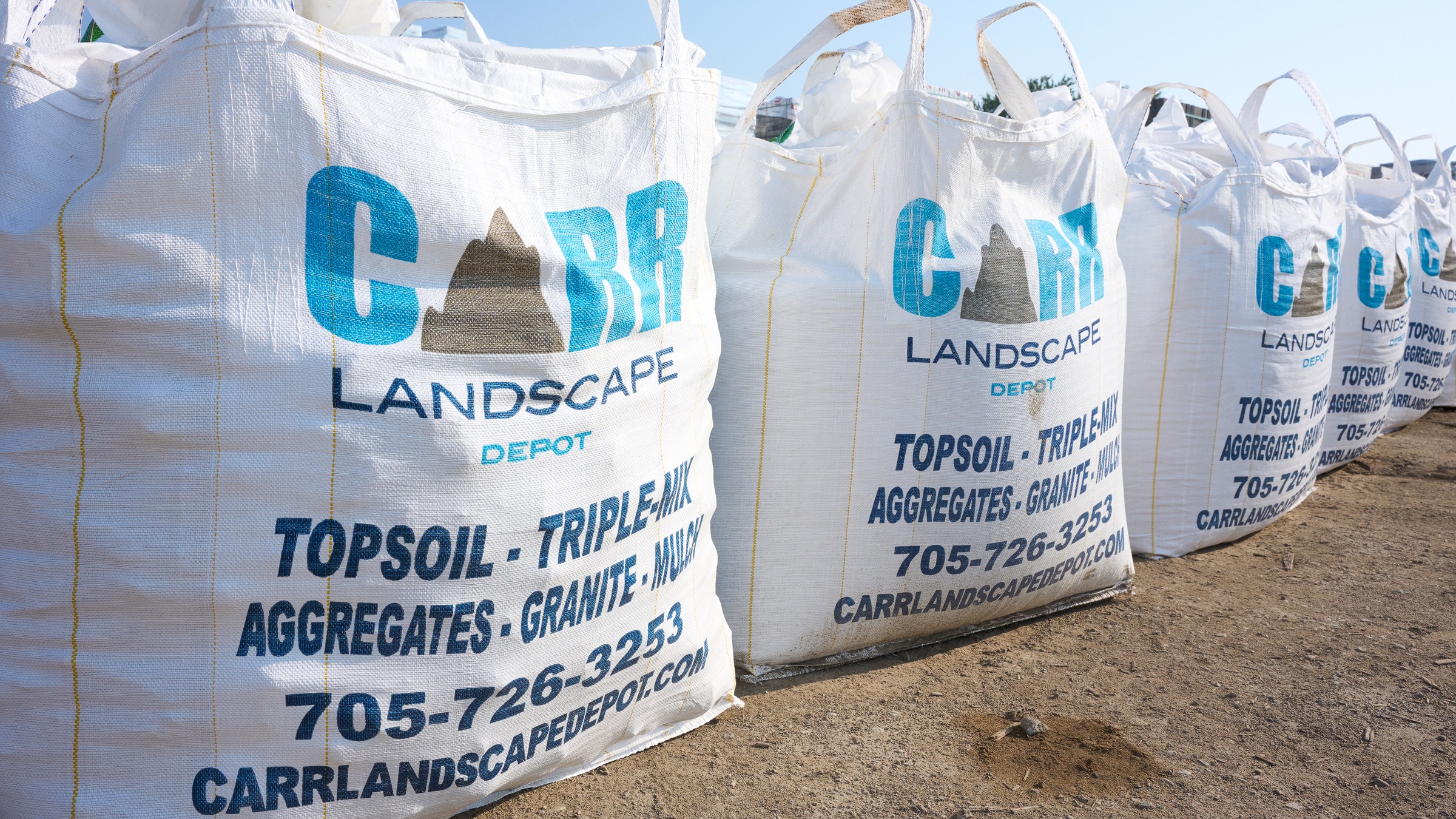 Large white bags filled with landscape materials, labeled "CARR Landscape Depot." The bags are neatly lined up outdoors on a dirt surface.