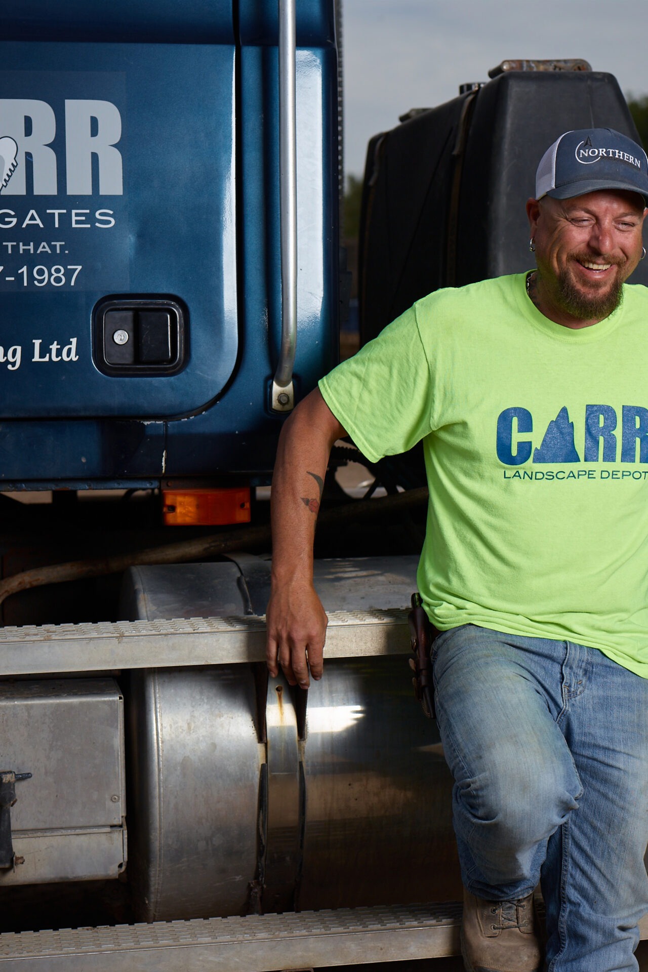 A person in a neon green shirt stands beside a blue truck, smiling. The shirt reads "CARR Landscape Depot" and the truck displays "CARR."