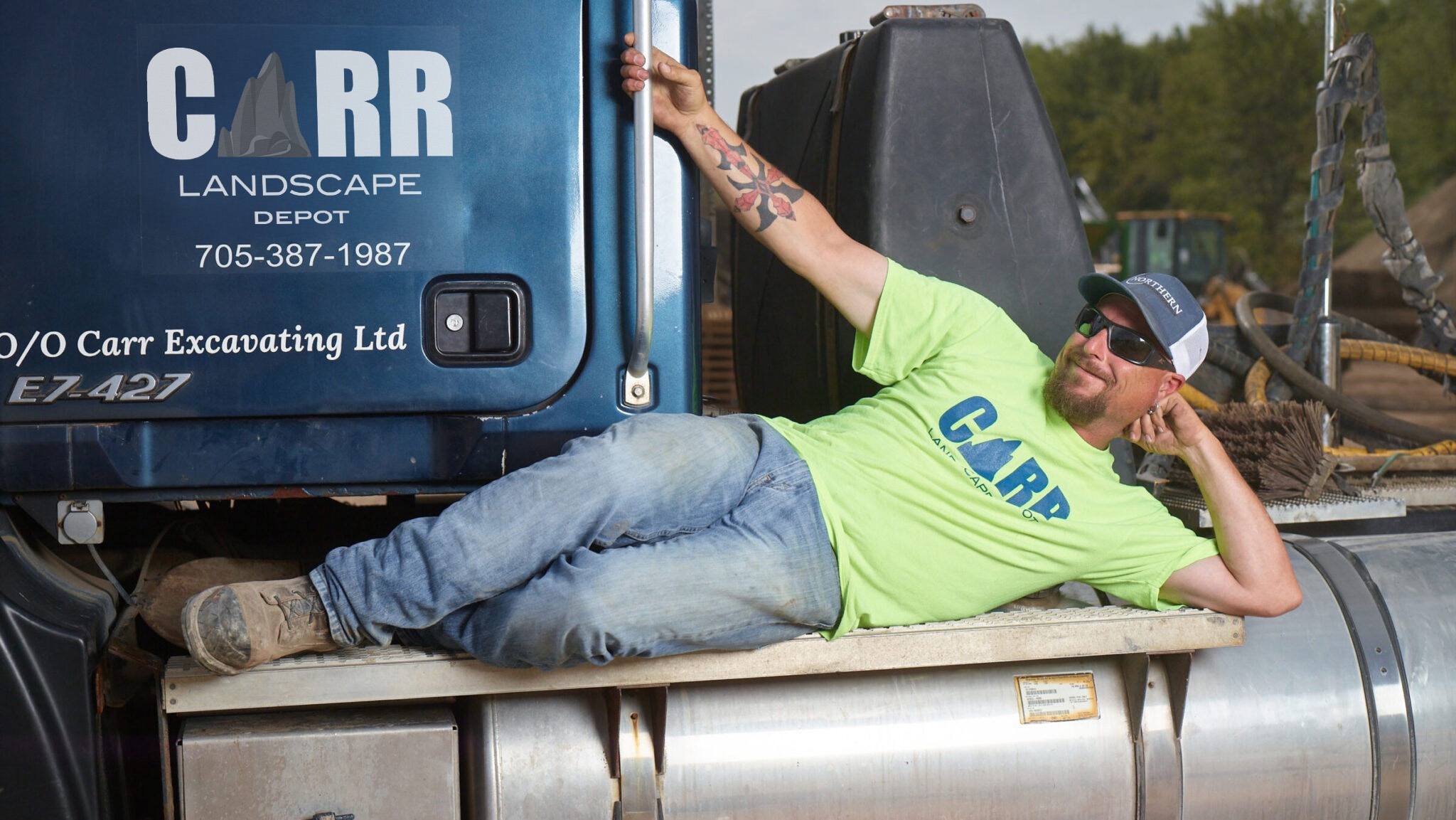 A person in a bright shirt lounges on a large vehicle labeled "CARR Landscape Depot" outdoors, surrounded by industrial equipment and greenery.