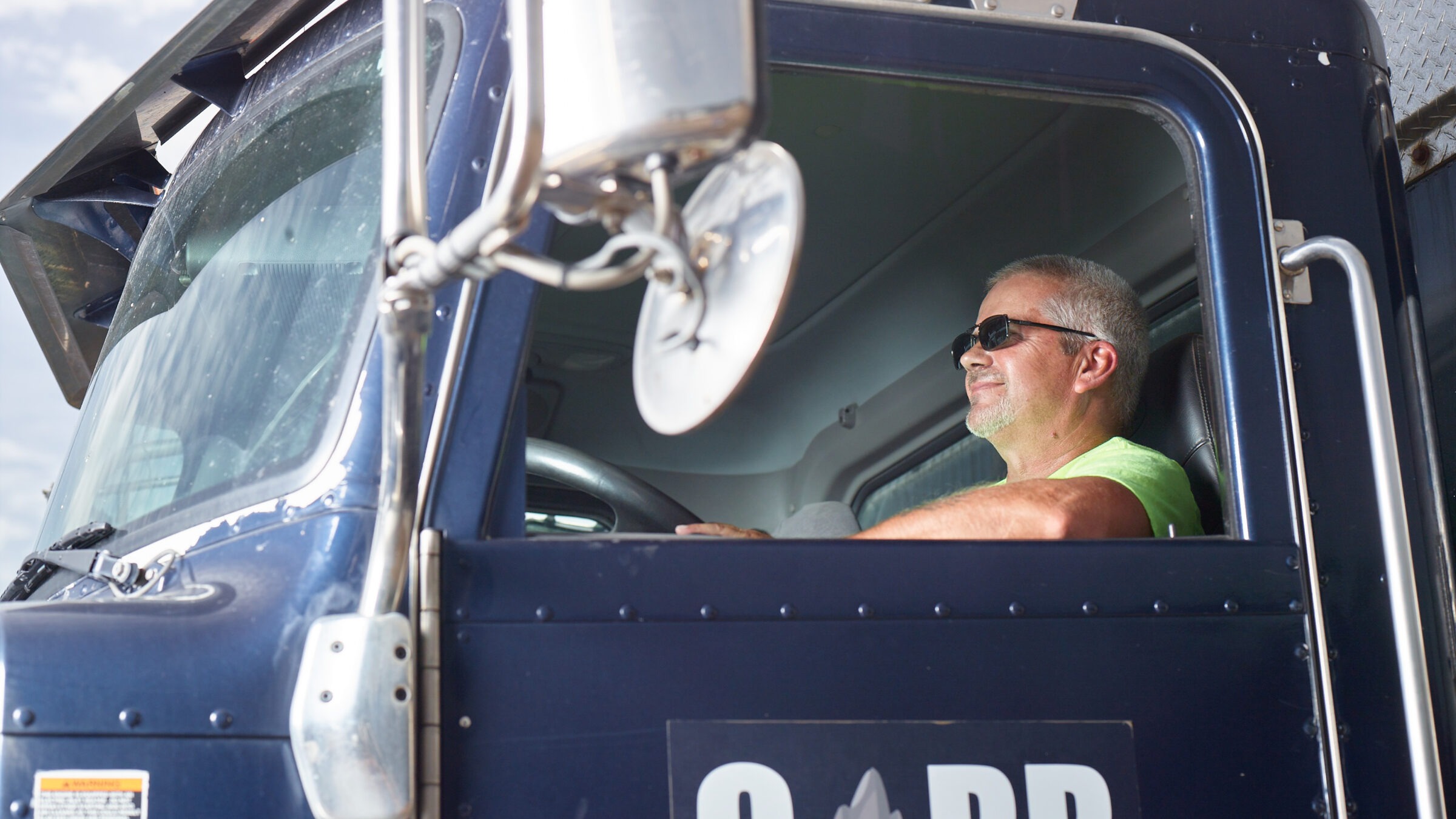 A person wearing sunglasses drives a blue truck with a visible logo, looking content through the driver's side window.