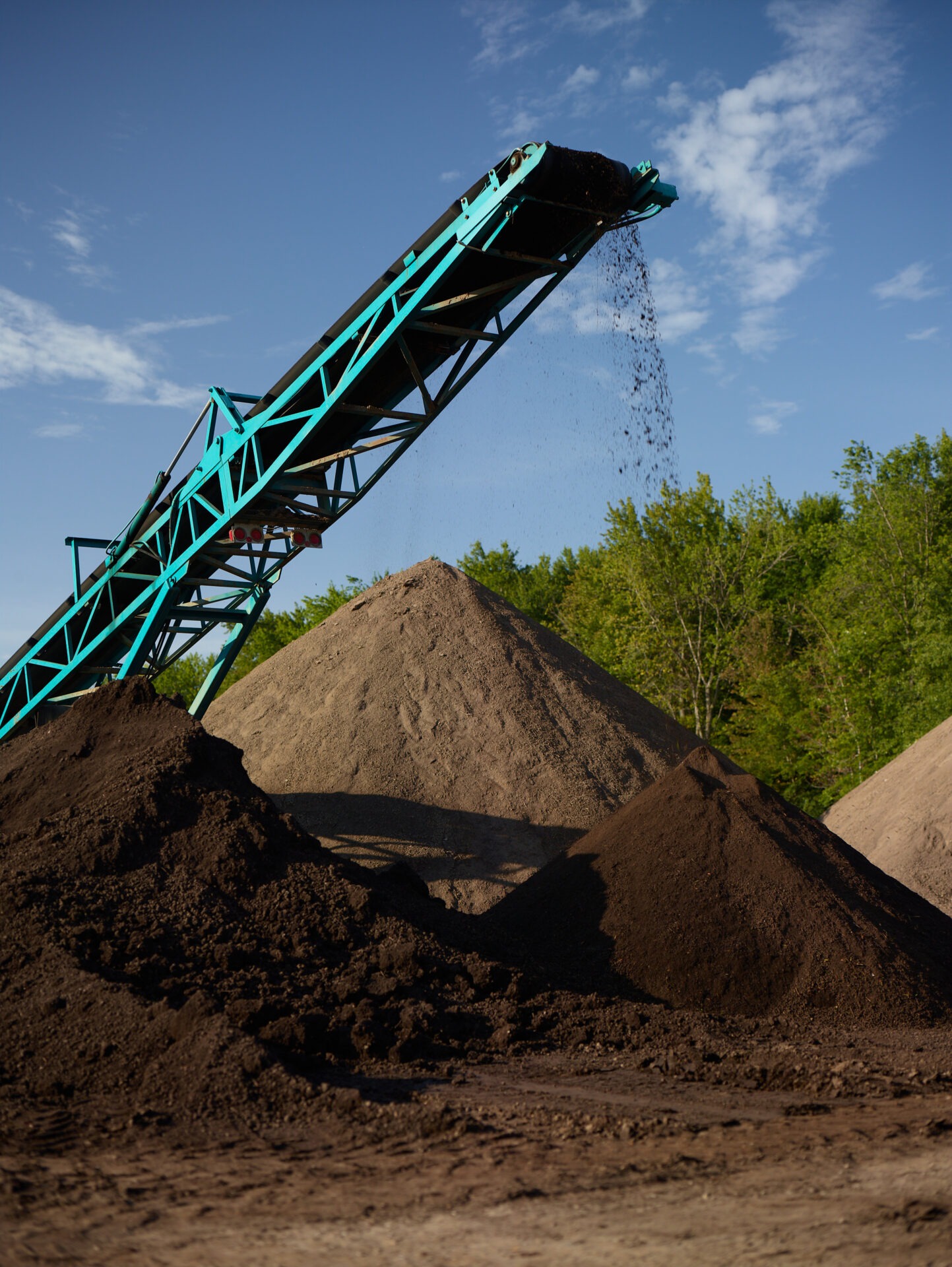 A conveyor belt deposits soil onto large mounds. Background features a clear sky and trees, suggesting a rural or industrial setting.