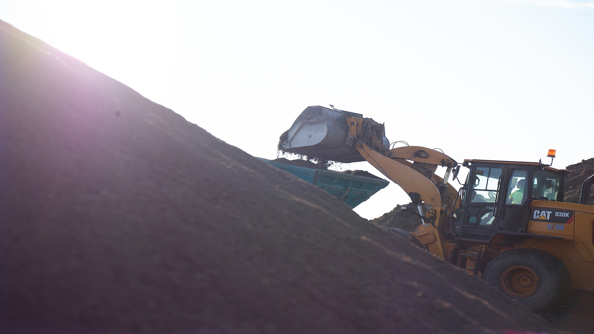 A large excavator moves dirt on a construction site under sunlight. The scene shows industrial equipment in action against a bright sky.