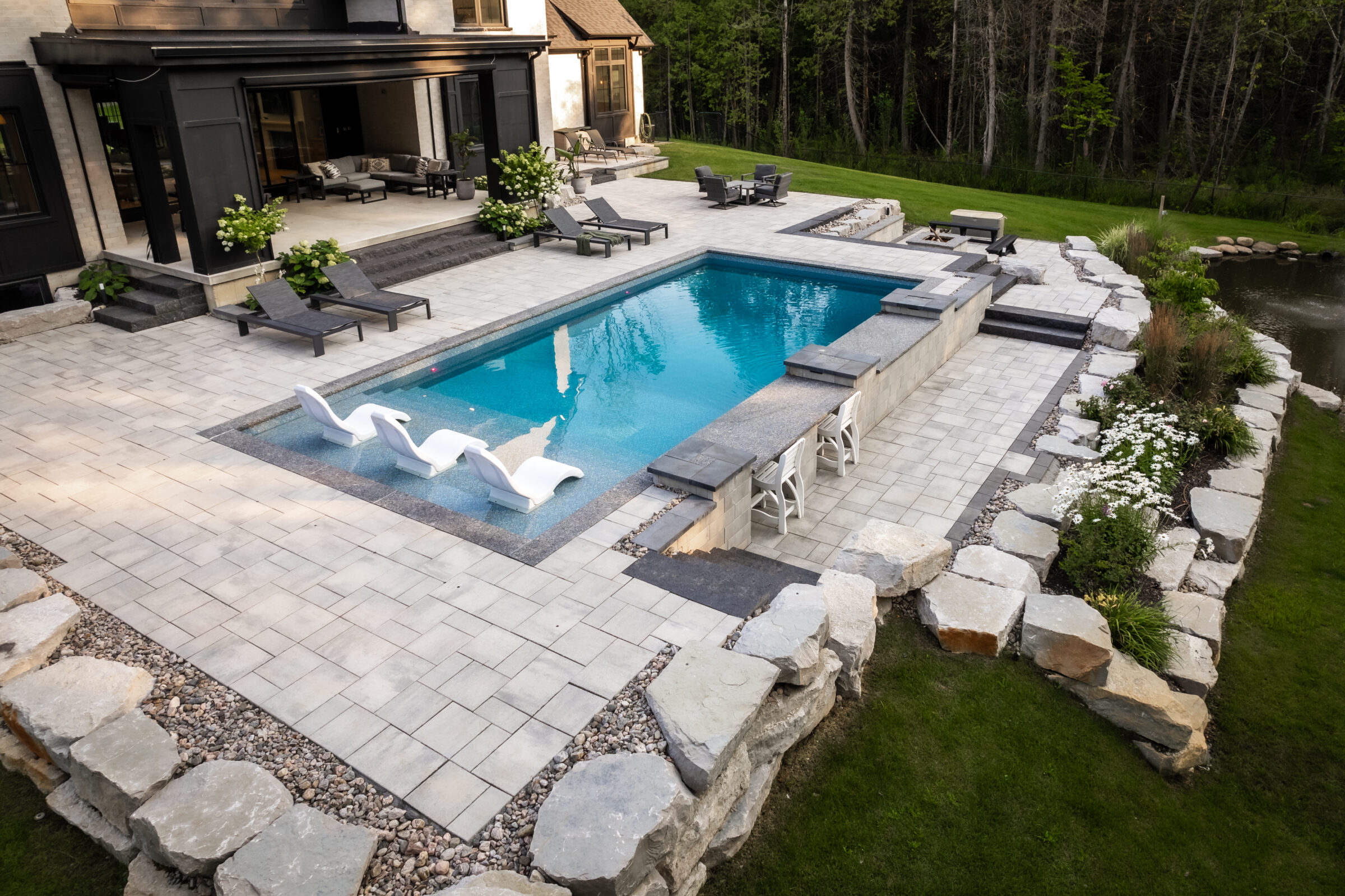 A modern outdoor space features a rectangular pool, lounge chairs, and landscaped garden. The area is surrounded by stone paving and natural greenery.