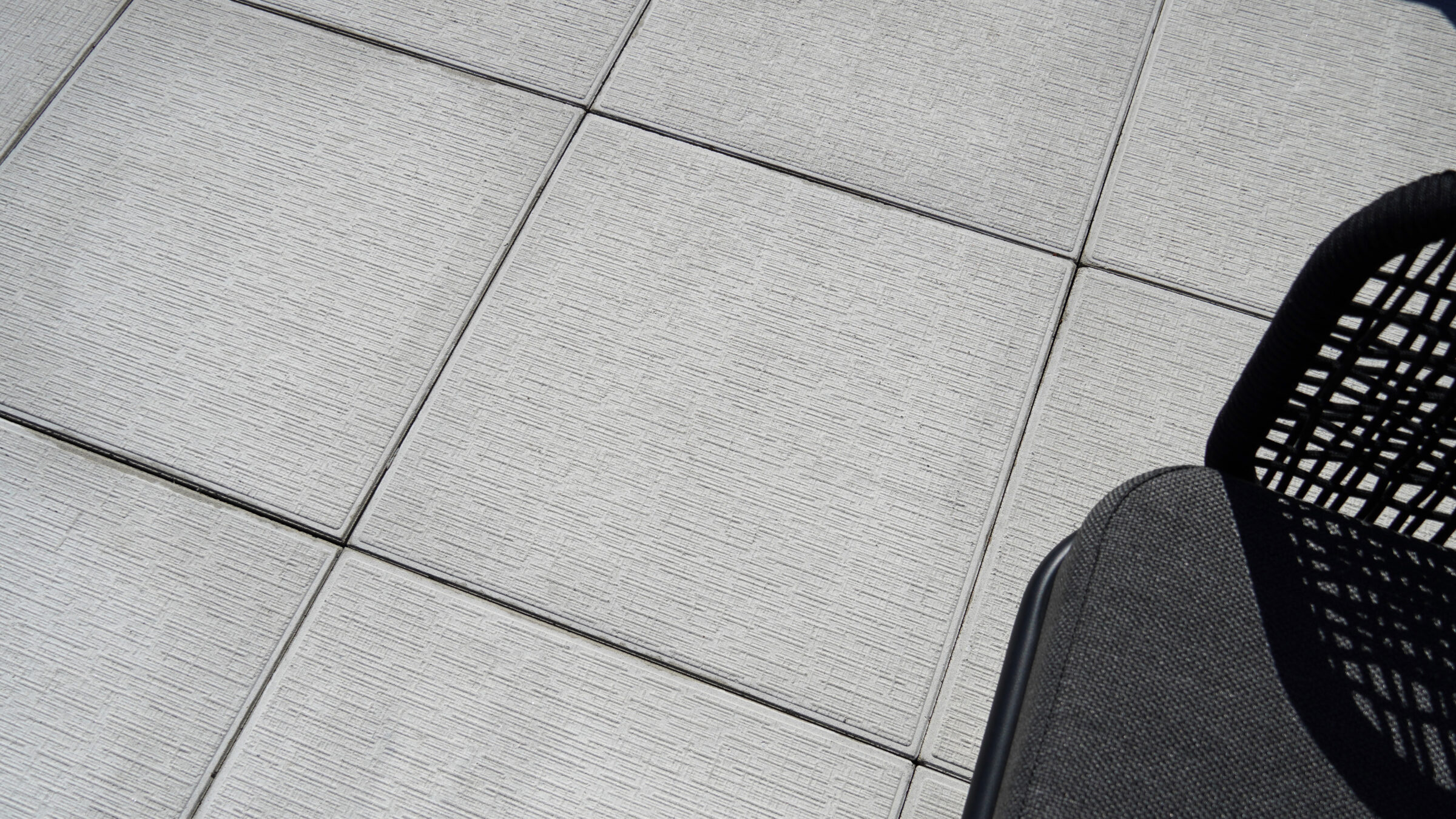 A black mesh chair sits atop light gray tiled flooring, creating a minimalist outdoor setting with clean lines and modern design elements.