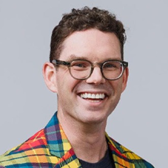 A person smiles, wearing a colorful plaid blazer and brown pants, against a simple gray background, conveying a cheerful, casual style.