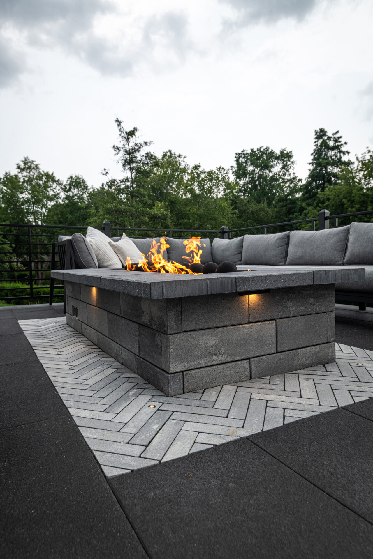 Outdoor patio with modern stone fire pit and gray sectional sofa. Surrounded by trees and overcast sky, creating a cozy, inviting atmosphere.