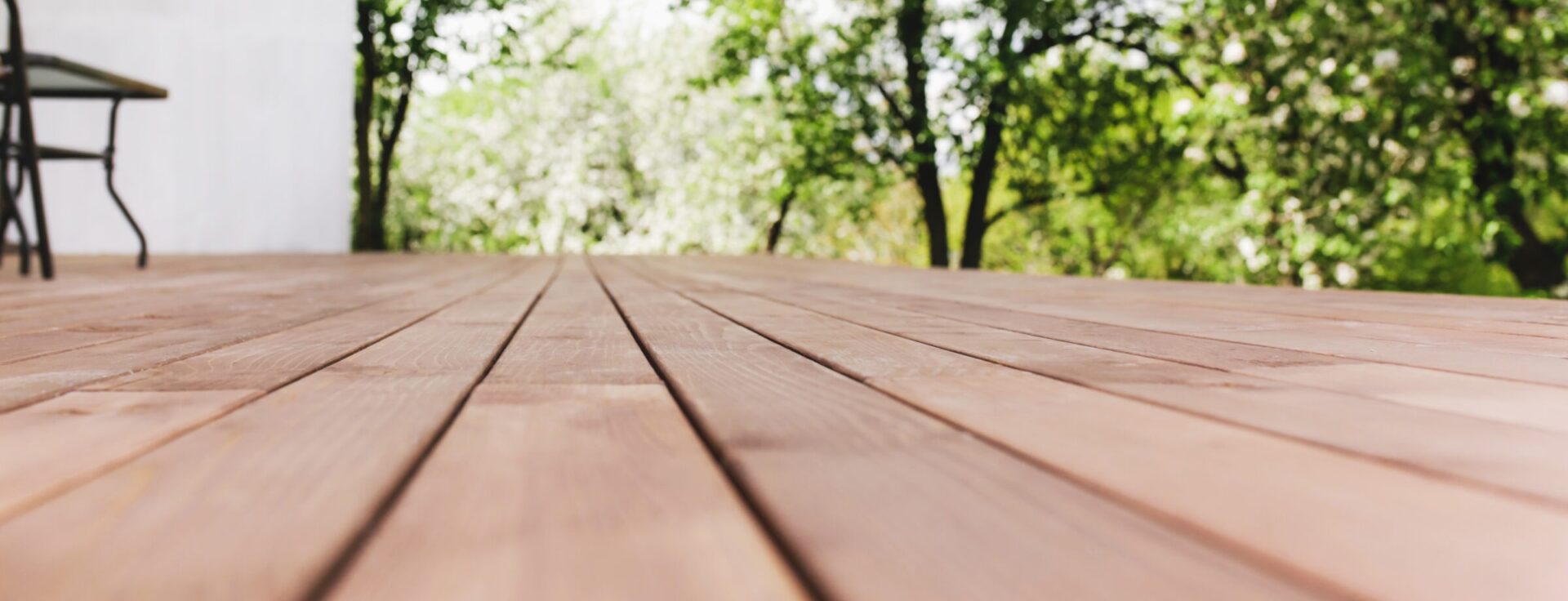 A wooden deck surrounded by green trees, adjacent to a white building, with outdoor furniture visible and sunlight filtering through the leaves.