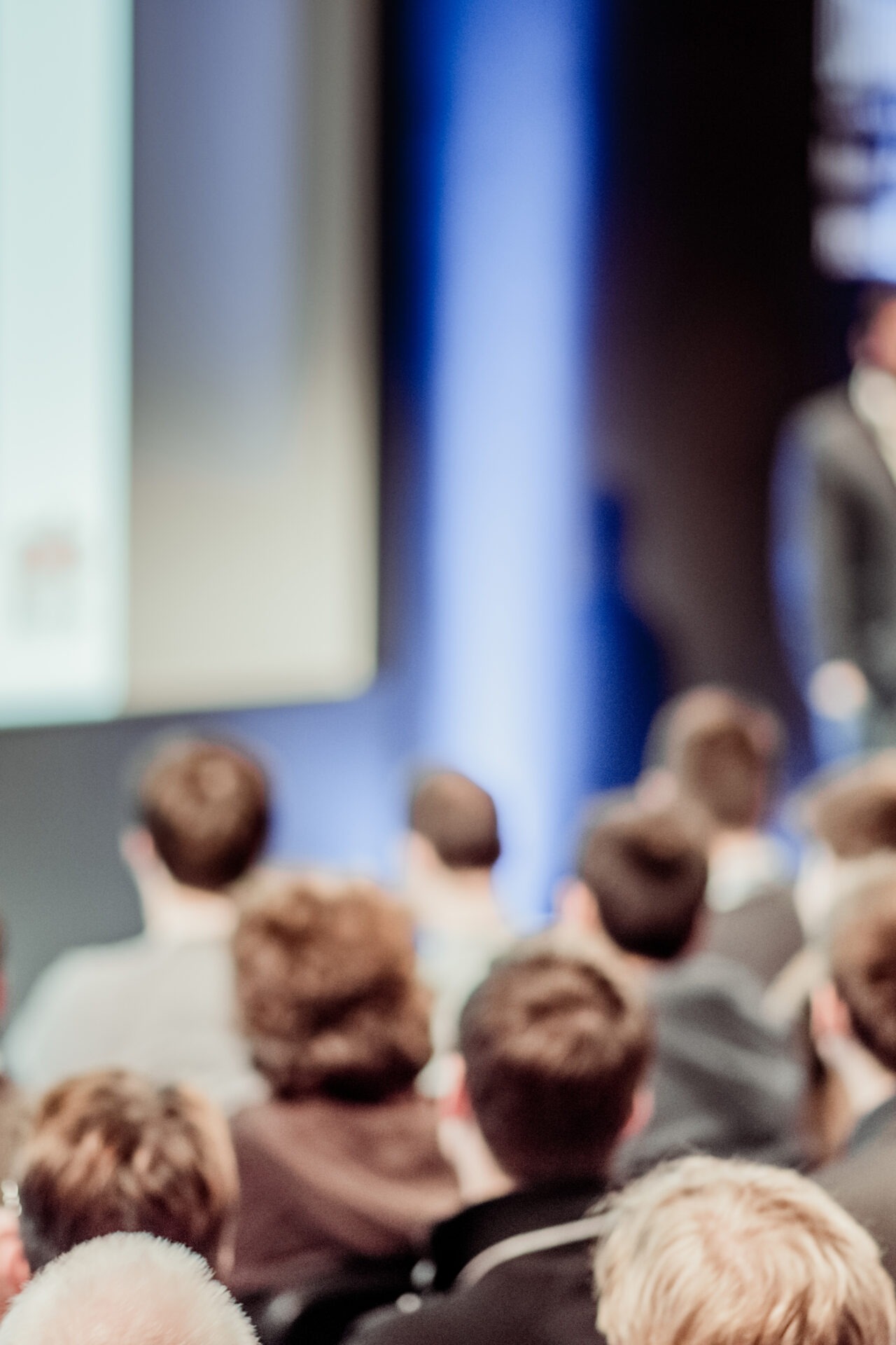 A group of people attentively watching a presentation in a conference room, with focus on the speaker near a projection screen.