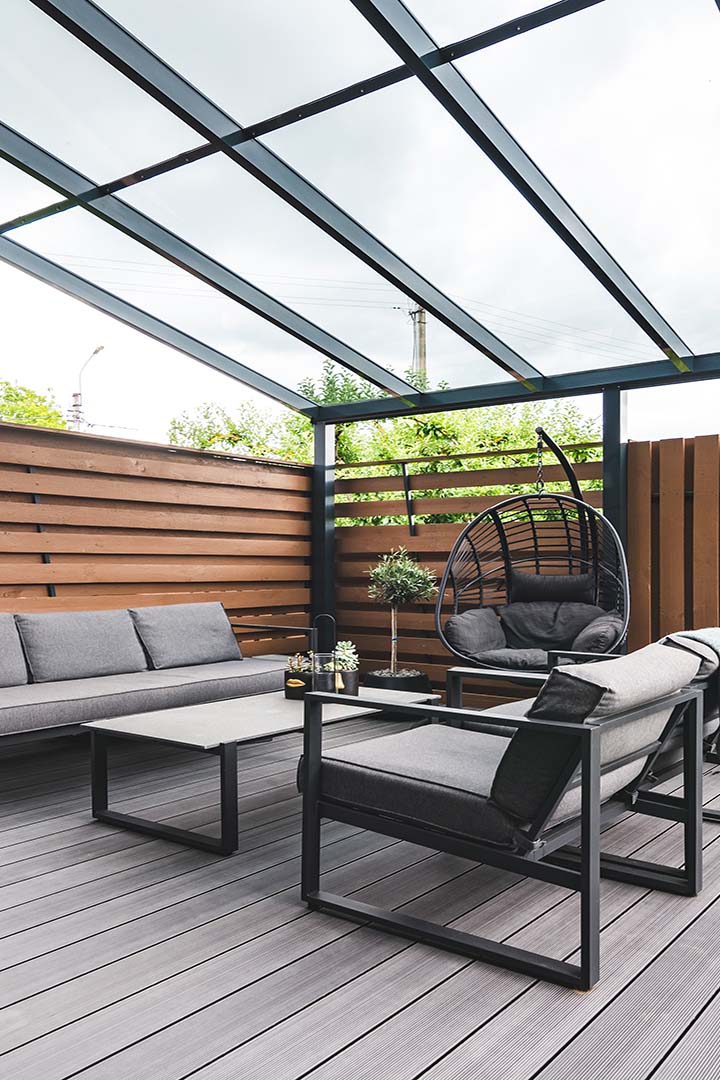 A modern outdoor patio with glass roof, featuring grey cushioned furniture, wooden fences, and potted plants, creating a cozy and stylish ambiance.