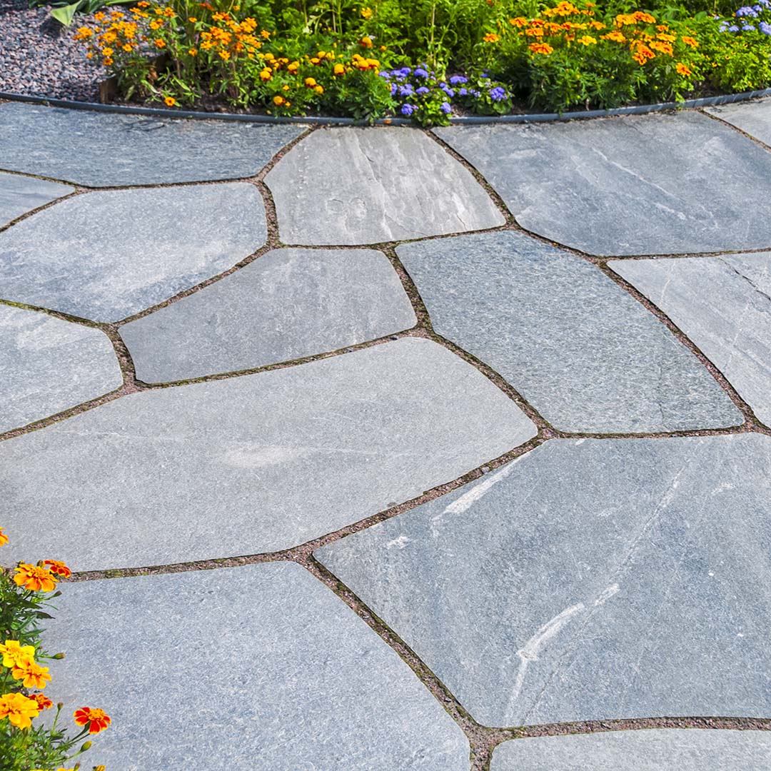 Stone path with irregular gray slabs bordered by green foliage and vibrant orange and purple flowers, creating a serene garden setting.