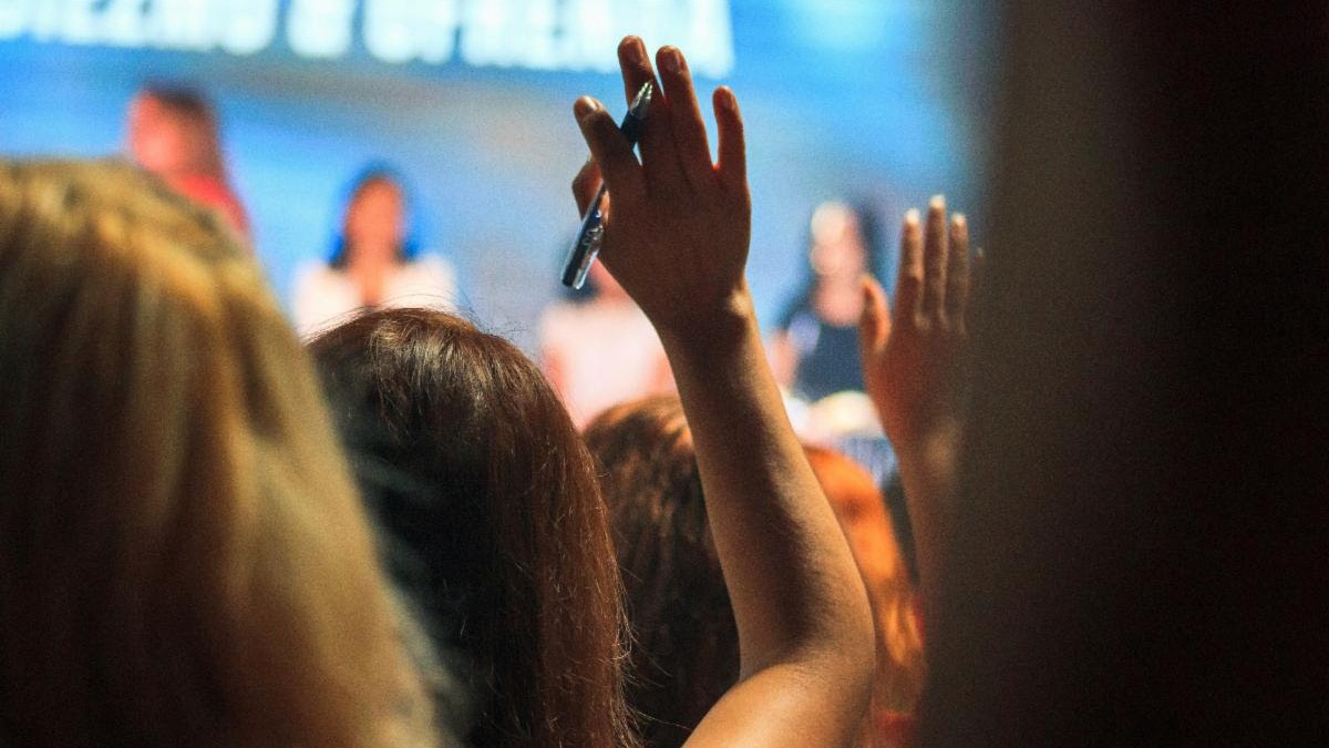 A group of people is gathered indoors, with hands raised, suggesting an interactive event or conference. Background slightly blurred, focusing on engagement.