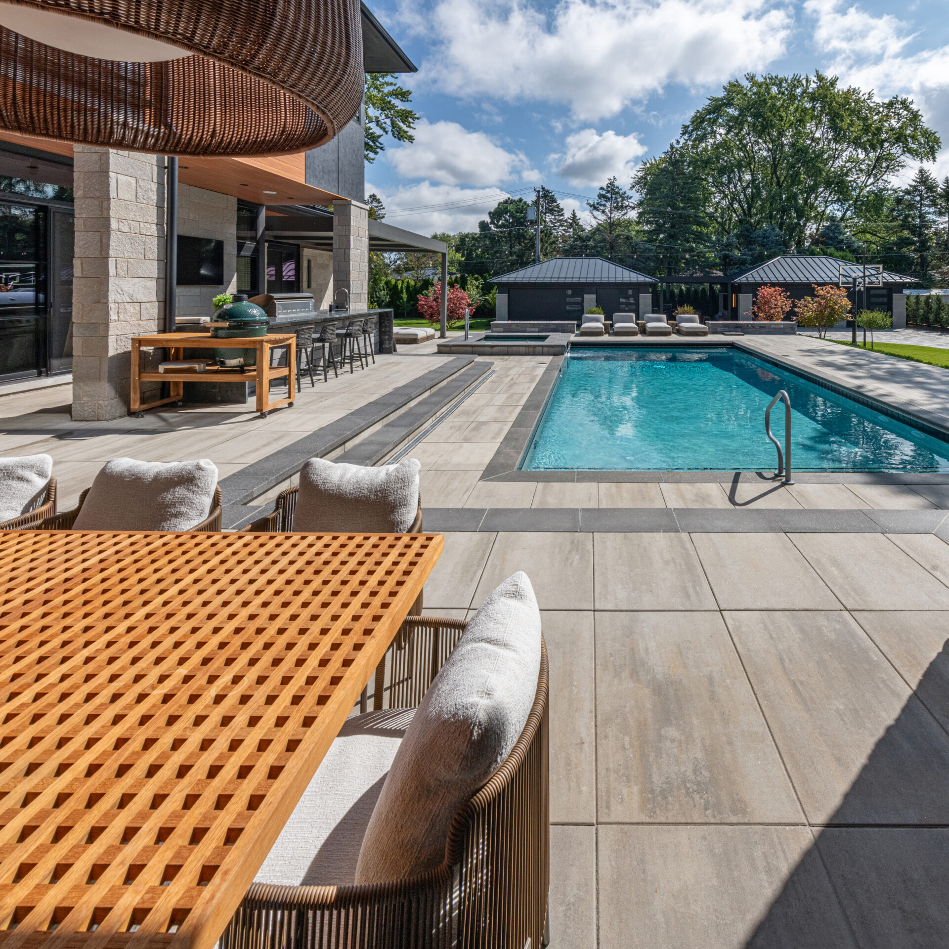 Modern outdoor living space features a large pool, lounge chairs, and a dining area. The setting is surrounded by lush trees under a blue sky.