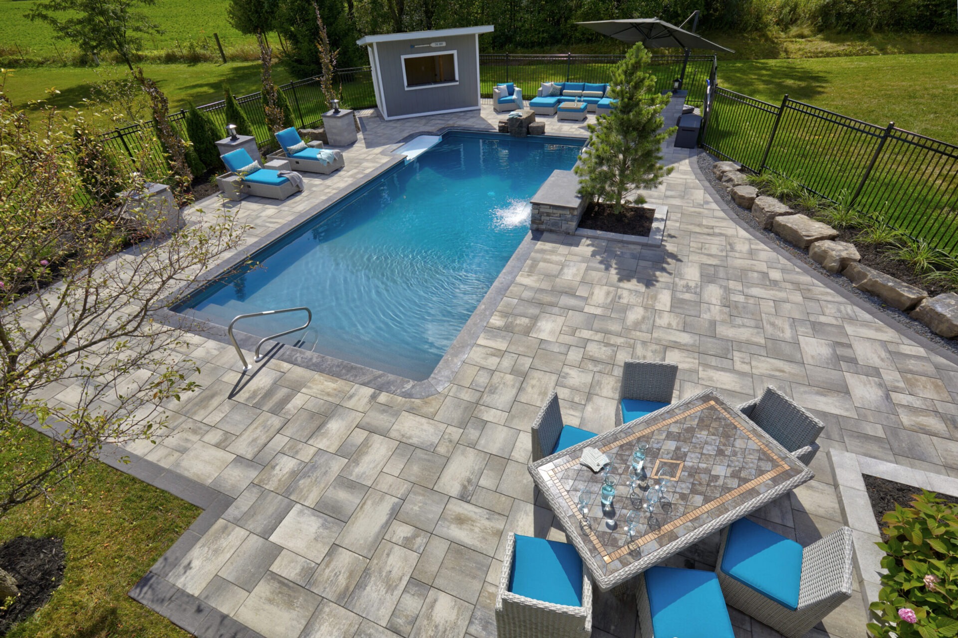 A backyard features a rectangular pool, lounge chairs, a small building, patio furniture, and greenery, enclosed by a metal fence.