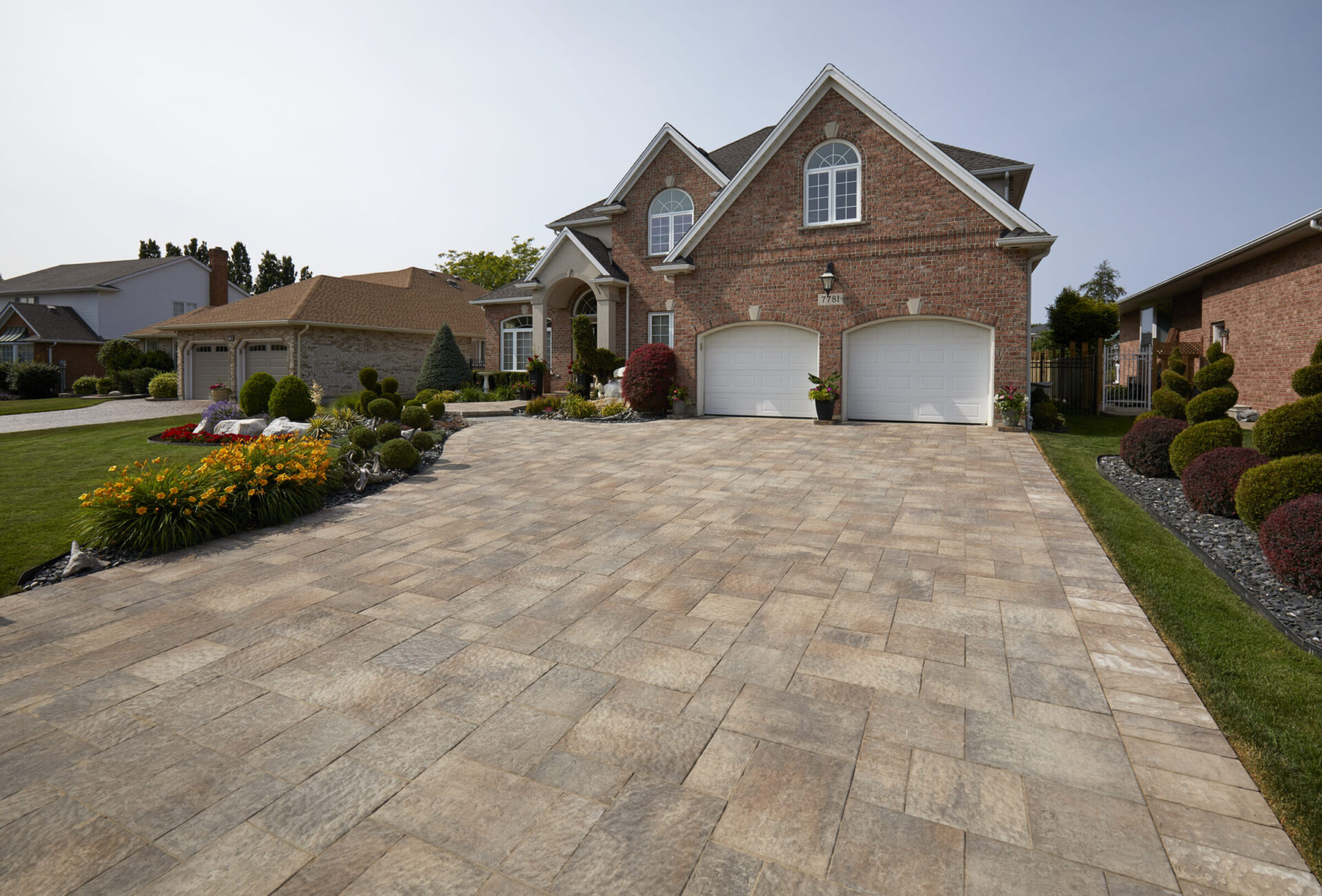 Brick house with a large paved driveway, surrounded by landscaped gardens and neatly trimmed bushes, under a clear sky. No people present.