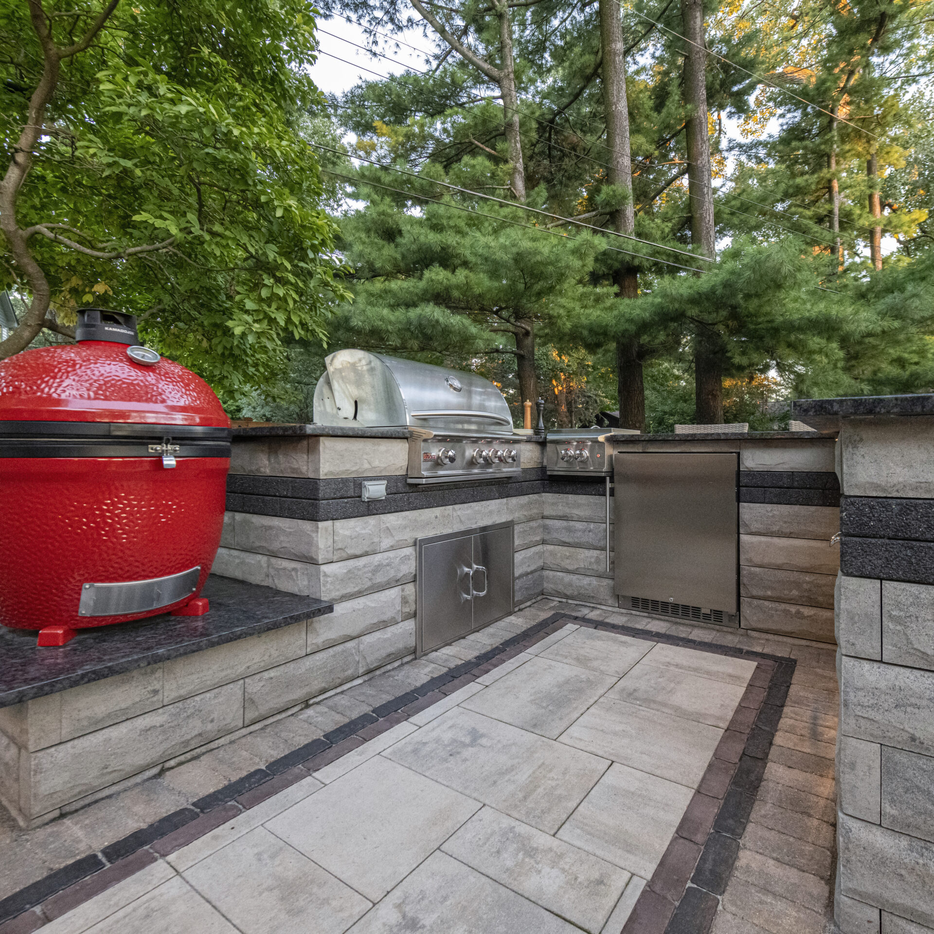 Outdoor kitchen features a red ceramic grill, stainless steel appliances, surrounded by trees and stonework, creating a serene backyard cooking area.