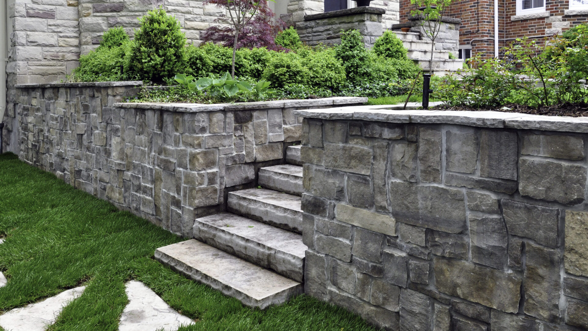 Stone steps with retaining walls bordered by green plants and grass. The scene is residential, featuring a charming combination of stonework and greenery.