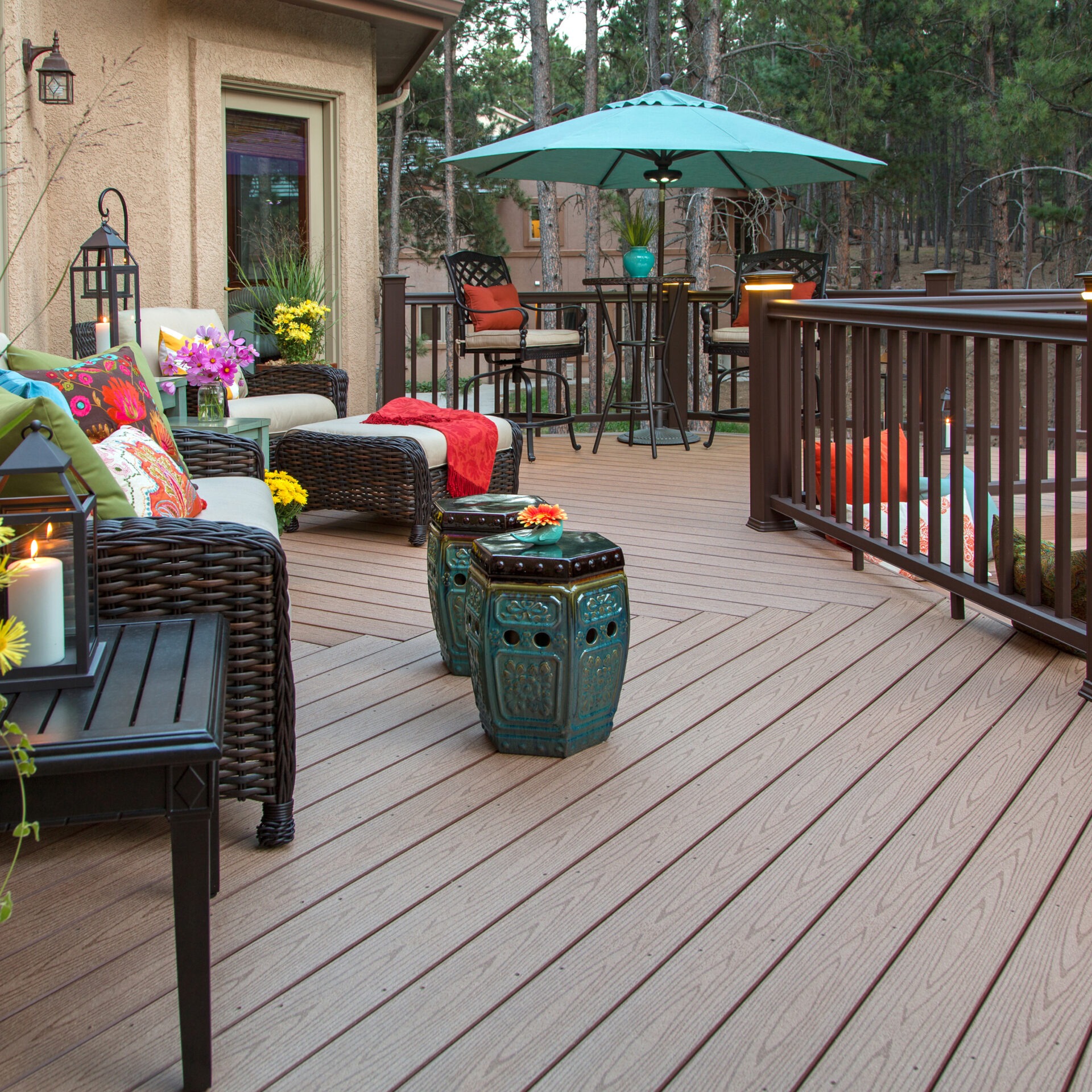 A cozy outdoor deck with wicker furniture, colorful cushions, and a teal umbrella, surrounded by trees. Lanterns and flowers add decorative touches.