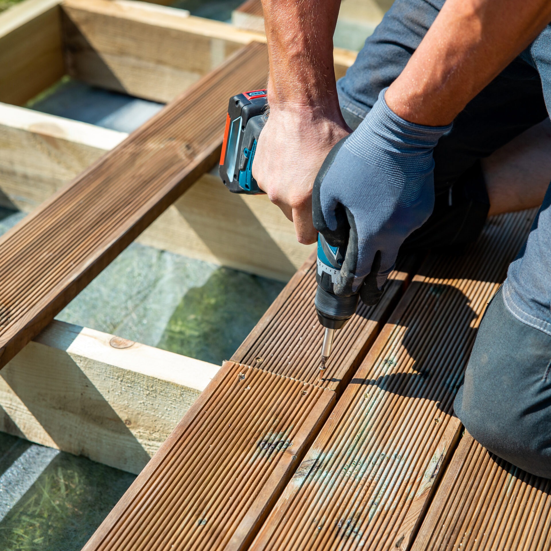 A person wearing gloves uses a power drill to screw wooden decking boards onto a frame, focusing on construction details and craftsmanship.