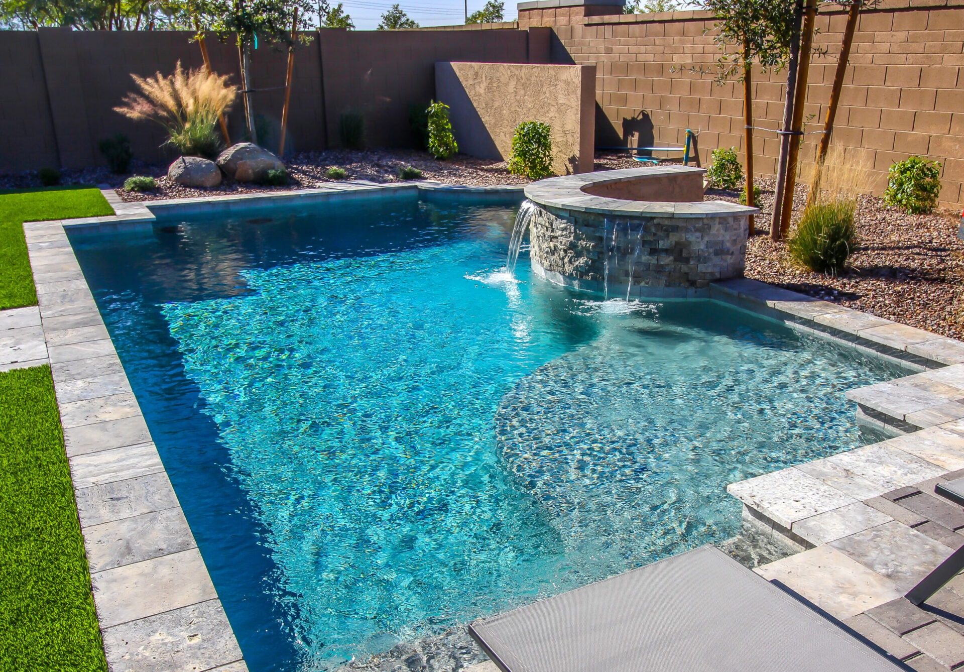 A backyard features a clear blue swimming pool with a small waterfall, surrounded by a concrete patio, grass, and privacy walls.