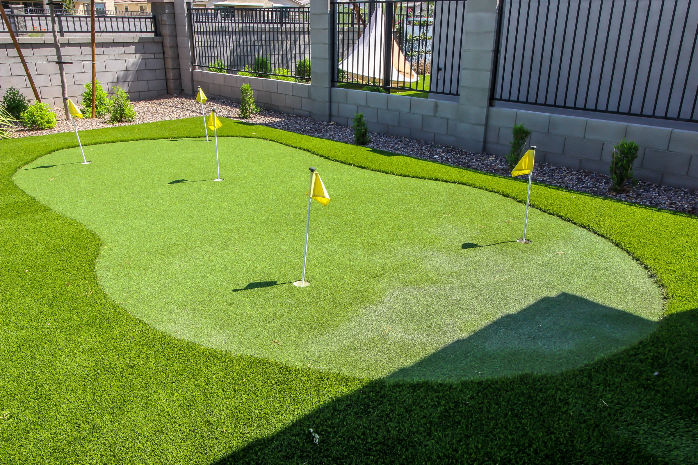 A small backyard putting green with four yellow-flagged holes, surrounded by artificial grass and bordered by a stone fence and metal railing.