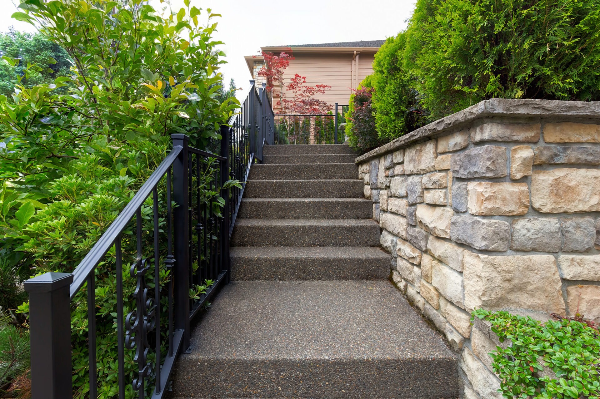 A stone staircase with black railings leads to a house, surrounded by greenery and a stone wall, creating a welcoming entrance.